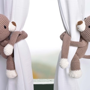 May include: Two brown crocheted monkey curtain tiebacks with white accents. The monkeys are holding onto white curtains.