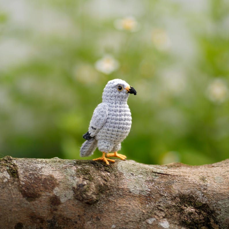 May include: A small, light gray crocheted bird figurine with a textured, yarn-stitched body. It features a black beak, dark eyes, and bright yellow feet. The bird is crafted with visible crochet stitches, showing a detailed, rounded shape.