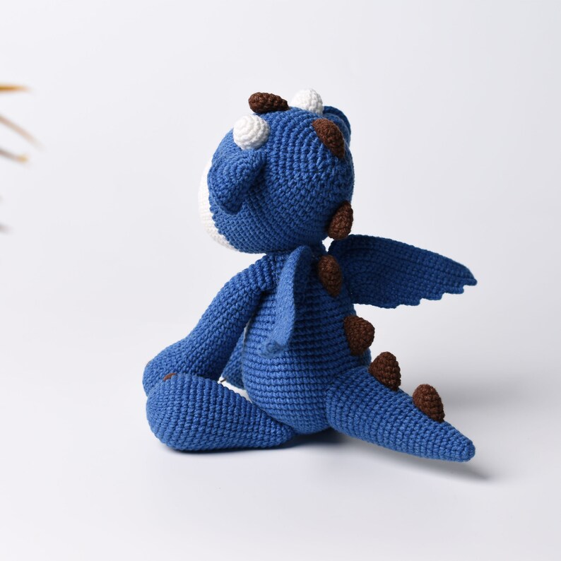 May include: A blue crocheted dragon toy with brown spots and white accents. The dragon is sitting with its wings spread out.