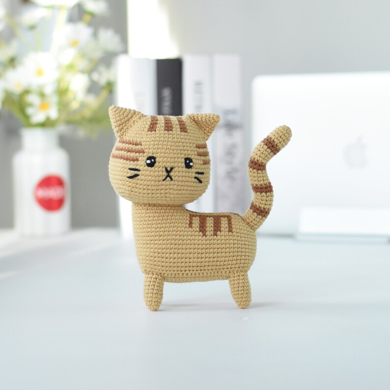 May include: A crocheted cat toy with brown stripes, a beige body, and a cute embroidered face.