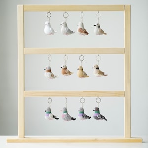 Peut inclure: Présentation de porte-clés oiseaux crochetés faits à la main, dans diverses couleurs, dont blanc, gris, marron et vert. Les porte-clés sont suspendus à un présentoir en bois. Chaque oiseau a un porte-clés en métal.