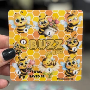 May include: A square card with a honeycomb pattern background and cartoon bees. The card features the text "BUZZ ALONG" and "TOTAL SAVED 25". Each bee has a number and is holding flowers or other items.