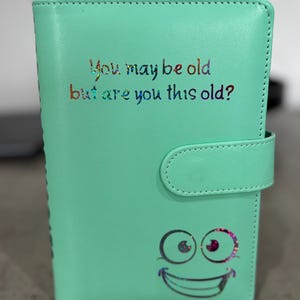 Może przedstawiać: Jasnozielony planer z napisem "You may be old but are you this old?" w kolorowych literach. Poniżej tekstu znajduje się grafika uśmiechniętej twarzy. Planer ma zapięcie na zatrzask.