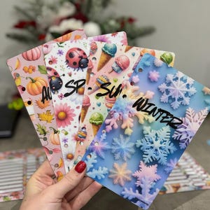 May include: A hand holds four seasonal cards. The cards are decorated with designs for Autumn, Spring, Summer, and Winter. The Autumn card has pumpkins and leaves. The Spring card has a ladybug and flowers. The Summer card has ice cream cones. The Winter card has snowflakes.
