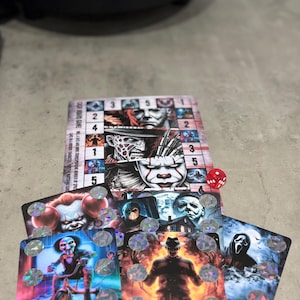 Könnte beinhalten: Horror-Spielkarten mit ikonischen Charakteren wie Freddy Krueger und Pennywise. Ein roter Würfel liegt neben den Karten. Das Spielbrett ist im Hintergrund sichtbar.