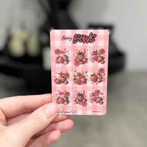 May include: A rectangular pink and white checkered savings challenge card titled Berry BOWS. The card features nine strawberry illustrations with pink bows and numbered circles for tracking progress. The text Total saved 25 is printed at the bottom.
