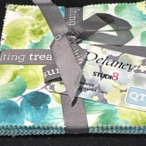 May include: A stack of square fabric pieces tied with a grey ribbon. The fabric features a floral pattern in shades of teal, light blue, and lime green. A paper tag attached to the ribbon displays the text Delaney, Studi8, and QT in black and red.