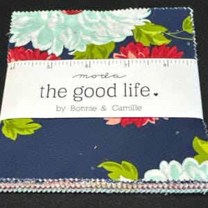 Puede incluir: Una pila de piezas de tela cuadradas con fondo azul marino y motivos florales en azul claro, rojo y verde. Una banda de papel blanco envuelve el centro, impresa con "moda the good life. by Bonnie & Camille" y una regla.