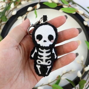 May include: A black and white plush skeleton keychain with a cute, cartoon-like face. The skeleton is wearing a black plush body with white embroidered bones.