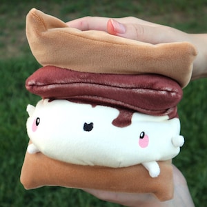 May include: A plush s'more toy. It features a brown graham cracker top and bottom, a dark brown chocolate layer, and a white marshmallow center with a cute face and small arms. The toy is held in a person's hand.