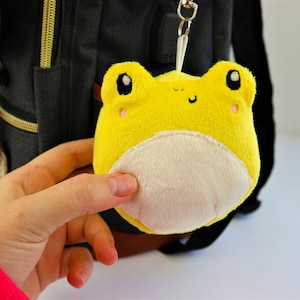 May include: A yellow plush frog keychain with a white belly and black eyes. The frog is attached to a black backpack.