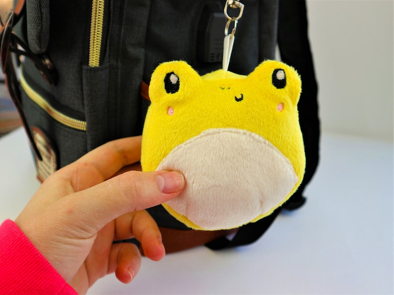 May include: A yellow plush frog keychain with a white belly and black eyes. The frog is attached to a black backpack.