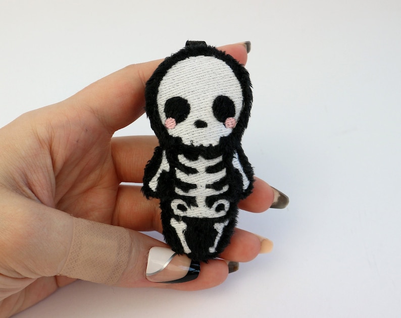 May include: A small plush skeleton toy with a black fuzzy body and a white embroidered skull with pink cheeks. The skeleton is wearing a black fuzzy outfit.