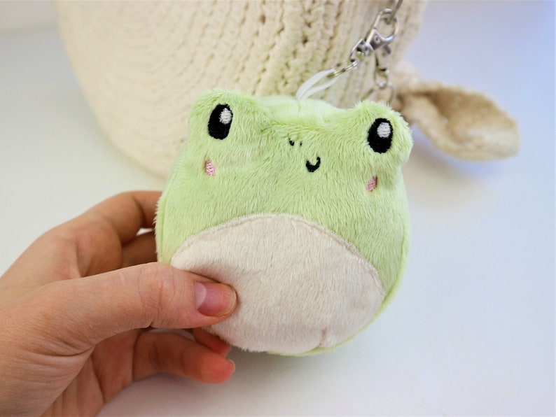 May include: A plush green frog keychain with black eyes and a white belly. The frog has a smiling face with pink cheeks.