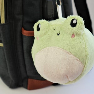 May include: A green plush frog keychain with a white belly and black eyes. The keychain is attached to a black backpack.