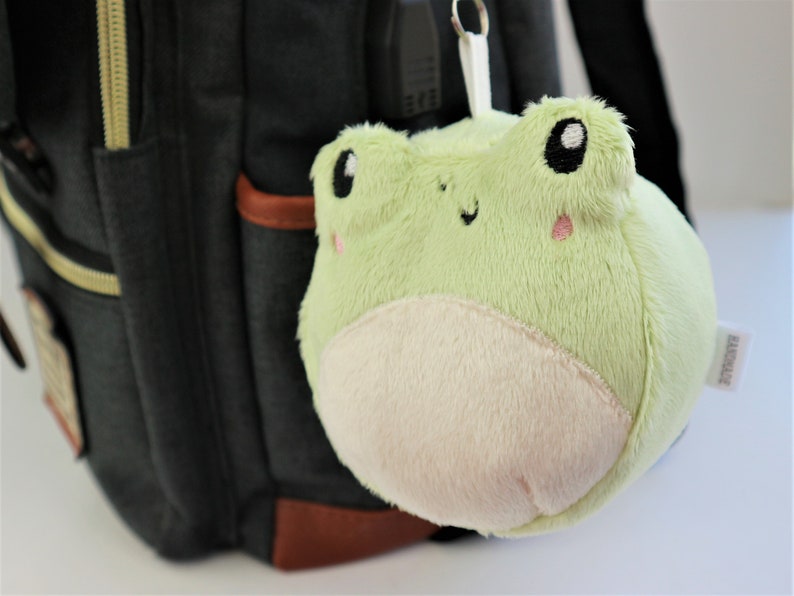 May include: A green plush frog keychain with a white belly and black eyes. The keychain is attached to a black backpack.