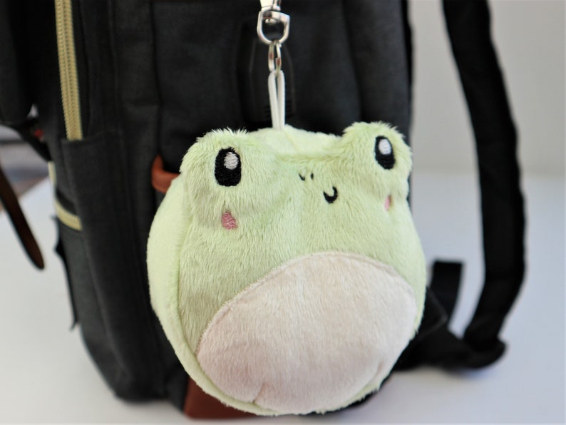 May include: A light green plush frog keychain with embroidered eyes, a pink nose, and a white belly. The frog is attached to a silver keychain.