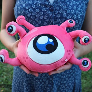 Può includere: Un giocattolo di peluche rosa con un grande occhio bianco con un'iride blu e una bocca sorridente. Il giocattolo ha quattro tentacoli rosa con occhi più piccoli su ogni tentacolo.