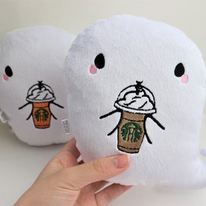 May include: Two white plush ghost-shaped cushions with embroidered faces and a Starbucks coffee cup with whipped cream. The ghost on the right has a tail.