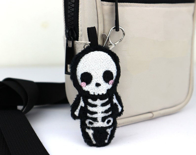 May include: A small plush skeleton keychain with black and white embroidery. The keychain is attached to a beige bag with a black zipper.