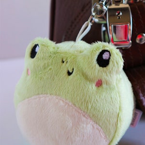 May include: A small, plush green frog keychain with white fur on its belly. The frog has black eyes with white pupils and pink cheeks. The keychain is attached to a silver metal ring.