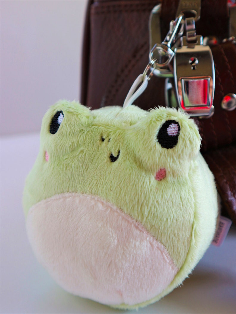 May include: A small, plush green frog keychain with white fur on its belly. The frog has black eyes with white pupils and pink cheeks. The keychain is attached to a silver metal ring.
