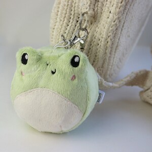 May include: A green plush frog keychain with black eyes and a white belly. The keychain is attached to a white crocheted bag.