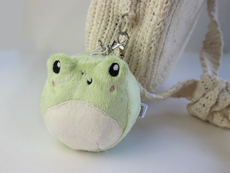 May include: A green plush frog keychain with black eyes and a white belly. The keychain is attached to a white crocheted bag.