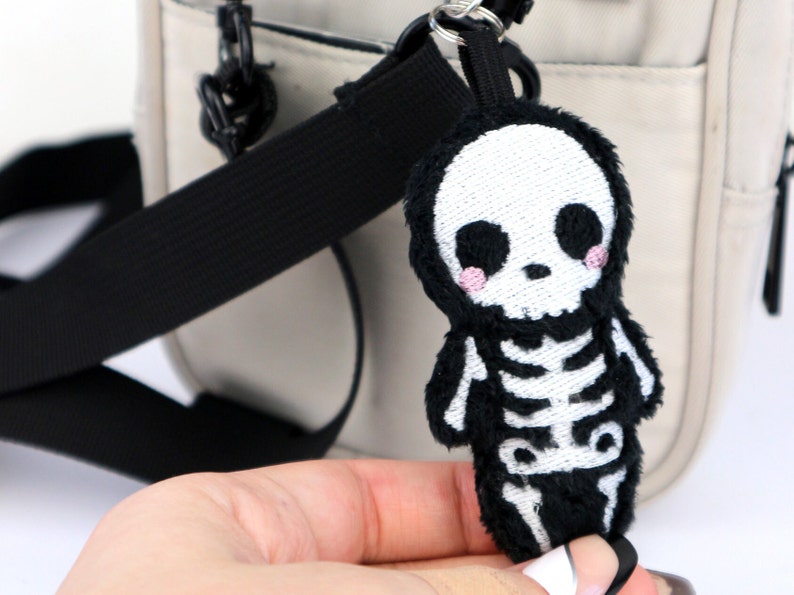 May include: A black plush skeleton keychain with a white embroidered skull and ribcage. The keychain is attached to a black and white striped strap.