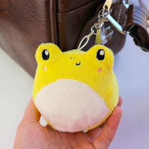 May include: A yellow plush frog keychain with white belly and black eyes. The frog is attached to a silver keychain.
