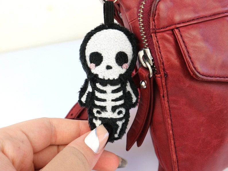 May include: A black and white skeleton plush keychain with a cute face and pink cheeks. The keychain is attached to a red leather bag.