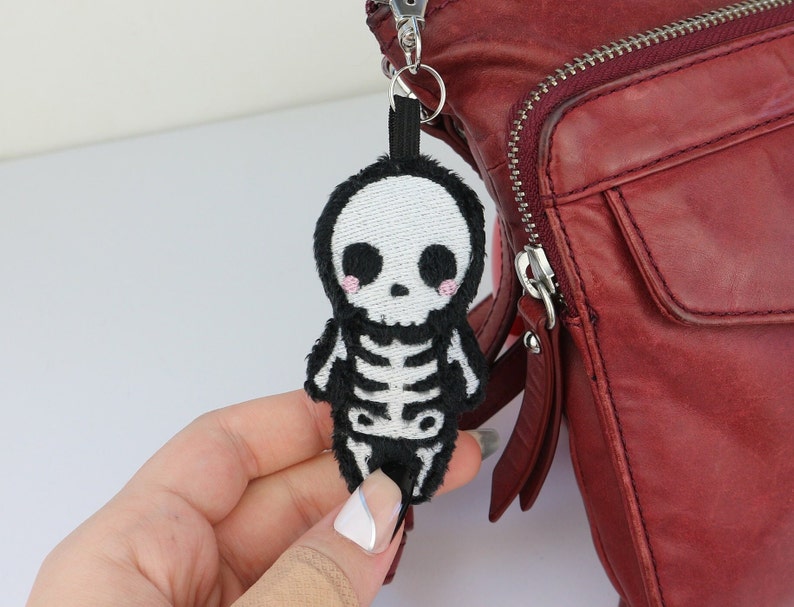 May include: A black and white skeleton plush keychain with pink cheeks. The keychain is attached to a red leather bag.