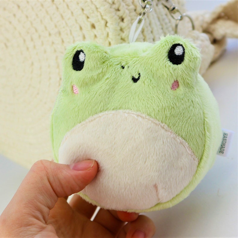 May include: A green plush frog keychain with a white belly and embroidered eyes and a smile. The keychain is handmade and has a tag that says "Handmade".