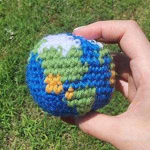 May include: A spherical crochet toy designed to resemble the planet Earth, featuring blue yarn for the oceans, green yarn for the landmasses, and white yarn for the polar ice caps, all worked in a dense, textured stitch pattern.