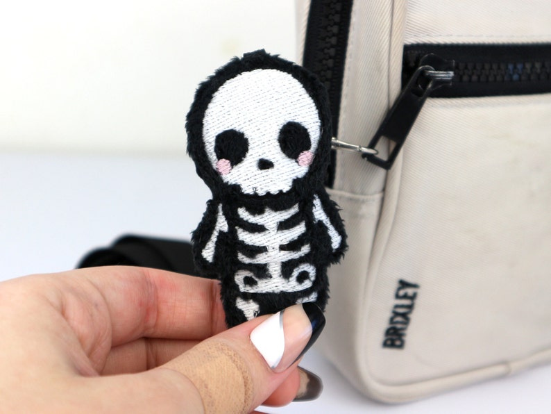 May include: A black and white plush skeleton keychain with a cute, cartoon-like face. The keychain is attached to a beige bag with the word "BROXLEY" printed on it.