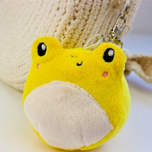 May include: A yellow plush frog keychain with a white belly and black eyes. The frog is smiling and has pink cheeks.