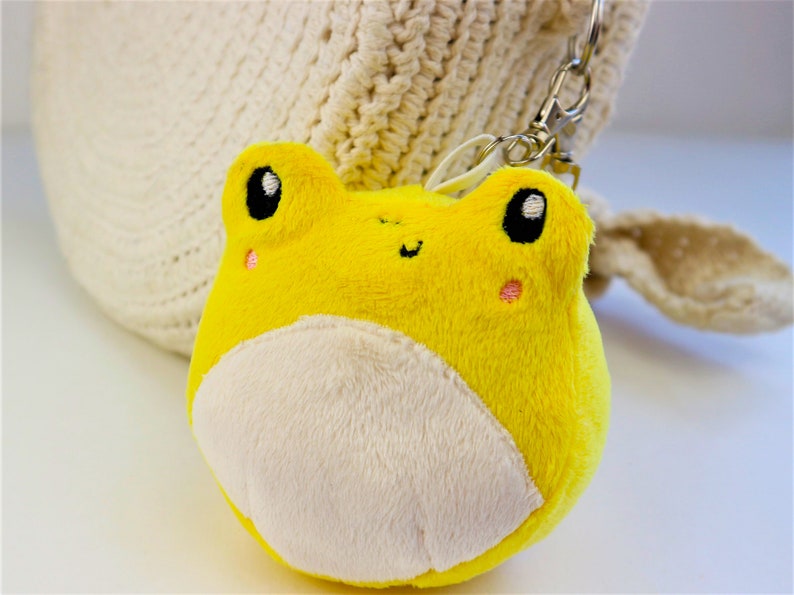 May include: A yellow plush frog keychain with a white belly and black eyes. The frog is smiling and has pink cheeks.