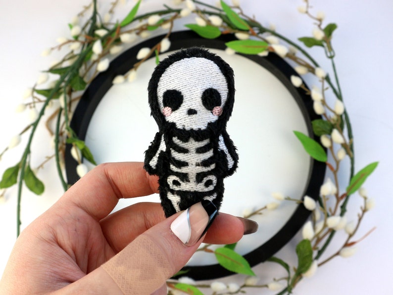 May include: A small, black and white embroidered plush toy shaped like a skeleton with a cute, cartoon-like face. The toy has a white skull with black eyes and a black ribcage.