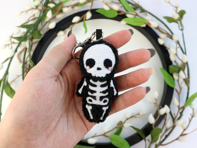 May include: A black and white plush skeleton keychain with a cute, cartoon-like face. The skeleton is wearing a black plush body with white embroidered bones.