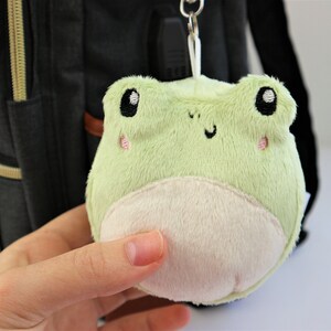 May include: A green plush frog keychain with embroidered eyes and a white belly. The frog is attached to a black backpack.