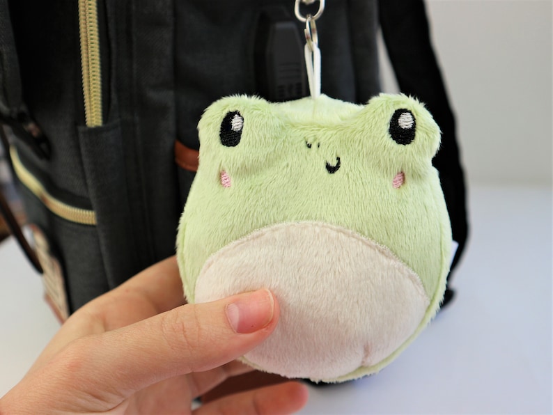 May include: A green plush frog keychain with embroidered eyes and a white belly. The frog is attached to a black backpack.