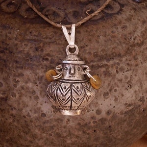 May include: A silver pendant necklace with an ornate, urn-shaped charm. The charm has a detailed pattern and two amber beads. It hangs from a thin cord. The pendant is displayed near a rustic, textured ceramic pot, evoking a vintage aesthetic.