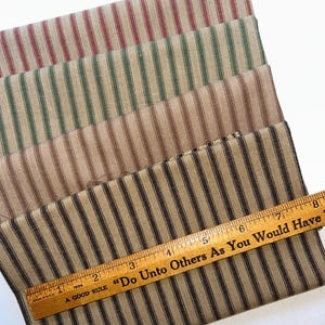 May include: Four fabric samples with vertical stripes in red, green, brown, and black. A wooden ruler with the text "Do Unto Others As You Would Have" is placed on the black and brown striped fabric. The ruler is marked with inches.