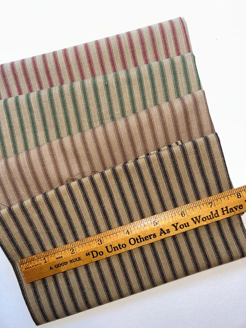 May include: Four fabric samples with vertical stripes in red, green, brown, and black. A wooden ruler with the text "Do Unto Others As You Would Have" is placed on the black and brown striped fabric. The ruler is marked with inches.
