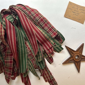 May include: A bundle of 50 red and green fabric strips in plaid and striped patterns, accompanied by a rusty metal star. A note reads: 50 CHRISTMAS RED N GREEN CHECK PLAID STRIPE HOMESPUN STRIPS 1/2"x18" & 1"x18". The fabric strips have frayed edges.