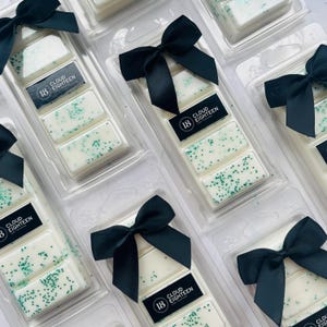 May include: Multiple clear plastic containers with black bows, each containing three white wax melts with green glitter. The wax melts are labelled "Cloud Eighteen".