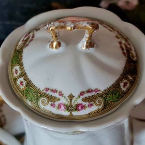 May include: Close-up of a white porcelain teapot with a scalloped lid. The lid features a floral design with pink roses, green accents, and gold trim. The handle and spout are also trimmed with gold.