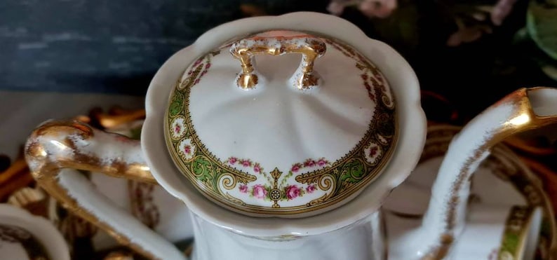 May include: Close-up of a white porcelain teapot with a scalloped lid. The lid features a floral design with pink roses, green accents, and gold trim. The handle and spout are also trimmed with gold.