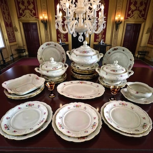 Könnte beinhalten: Ein formelles Essgeschirr-Set mit weißen Porzellangeschirr mit floralen Mustern und goldenen Verzierungen. Das Set umfasst Teller, Servierschüsseln und abgedeckte Gerichte, die auf einem dunklen Holztisch angeordnet sind. Eine große Kronleuchter hängt darüber.