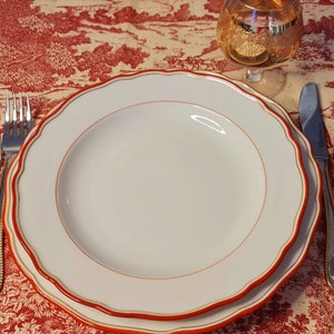 Puede incluir: Un plato blanco para cenar con un borde rojo y dorado, colocado sobre un mantel rojo y blanco con un patrón floral. Dos tenedores de plata y un cuchillo de plata se colocan a la derecha del plato. Una cuchara de plata se coloca a la derecha del cuchillo.